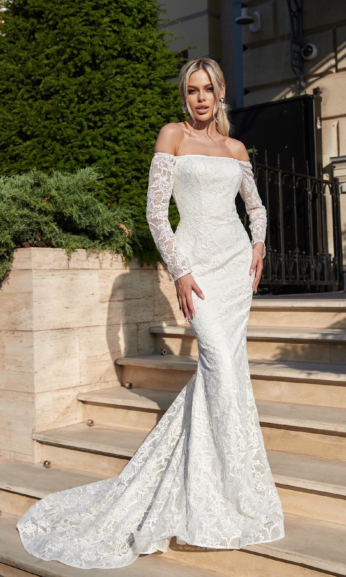A woman stands on outdoor steps wearing the Long Lace White Formal Gown: Tina Holly BB007—an off-the-shoulder, fitted, floor-length bridal dress with lace details and long sleeves.